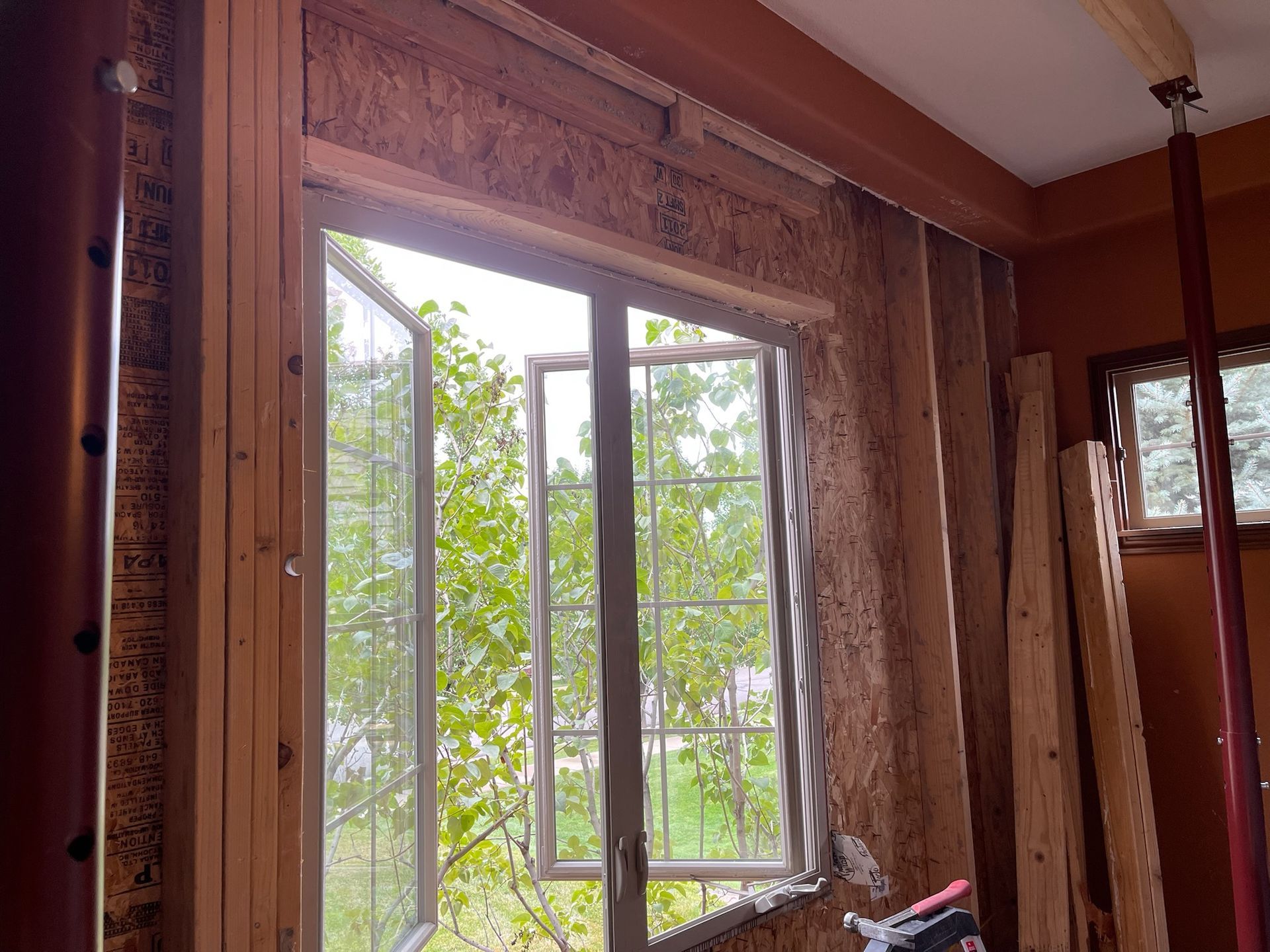 Interior view of an open window during home renovation. Wooden frame, exposed wall, trees outside, and supports visible.