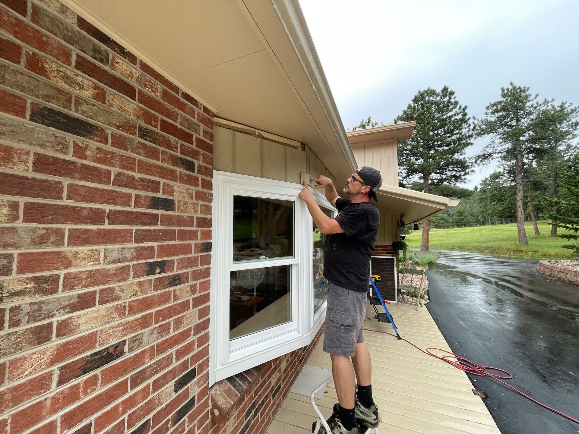 A person installing something near the window of a brick house under a tan soffit on a cloudy day.