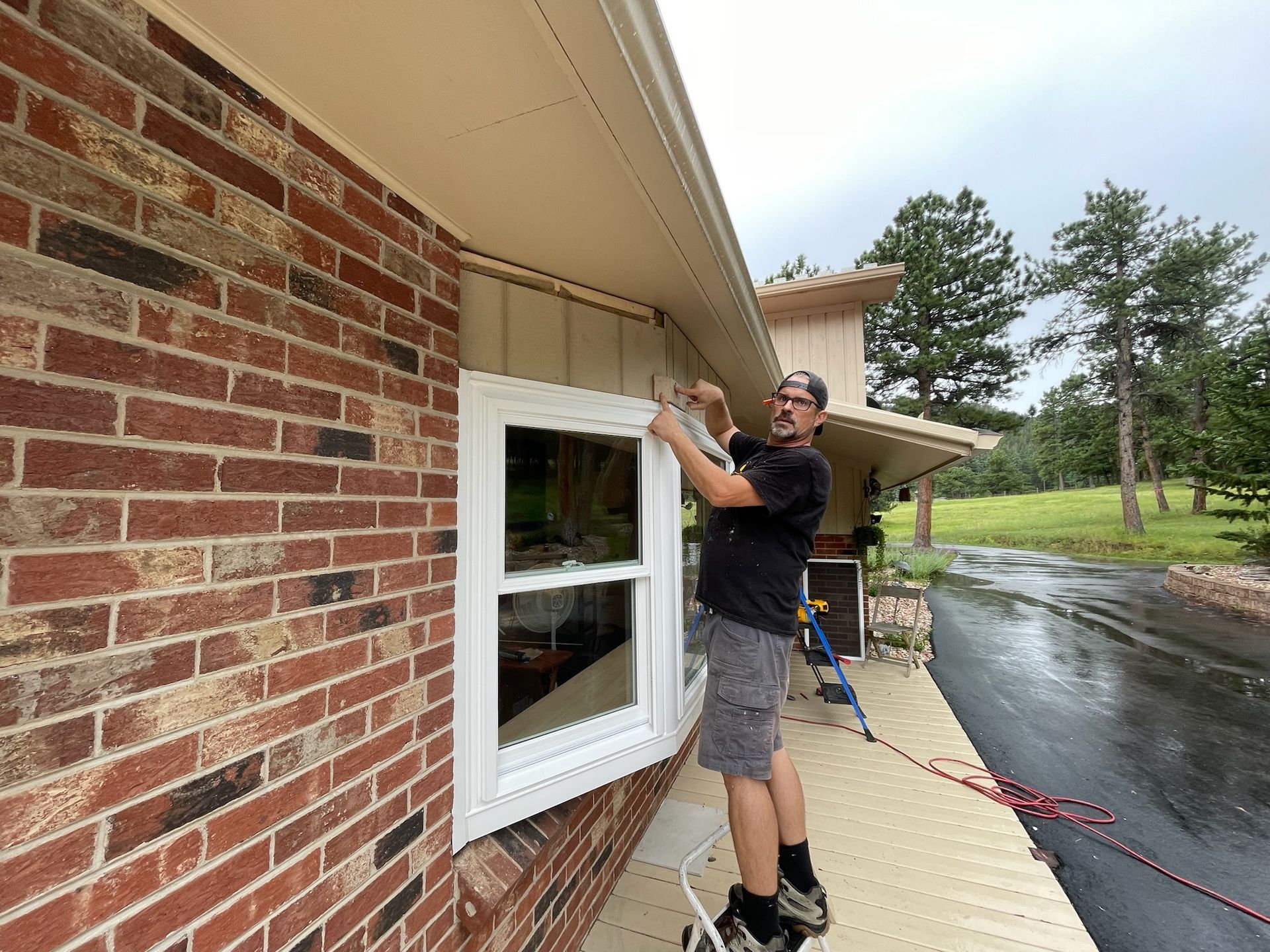 Man installing trim around a window on a brick house. He is standing on a ladder. Overcast day.