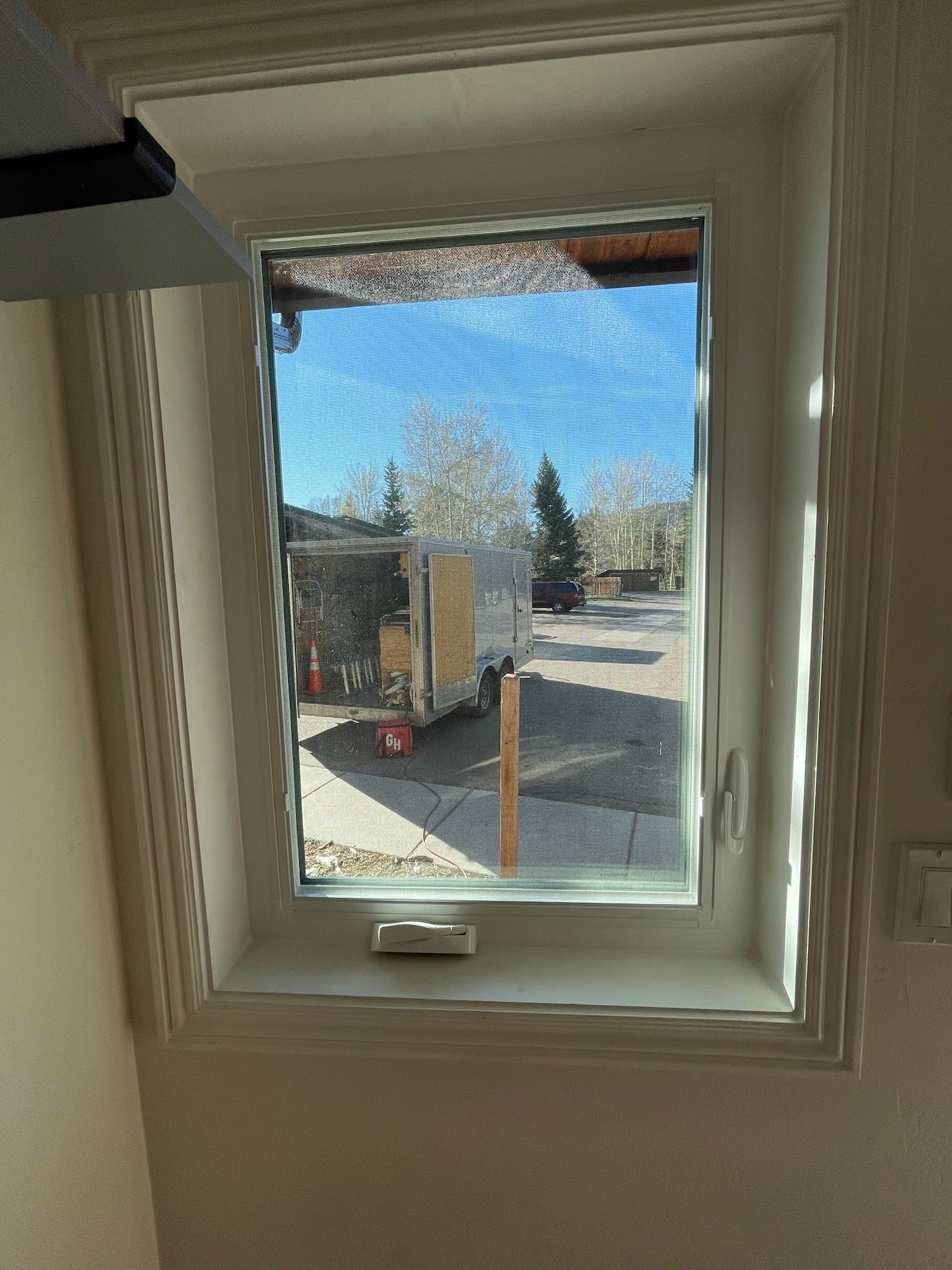 Window with screen, overlooking a street with a vehicle, trees, and buildings; bright sky.