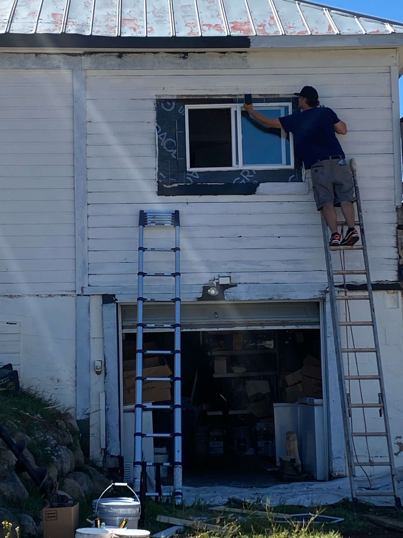 Person painting a window on a white building, standing on a ladder. A second ladder is nearby.