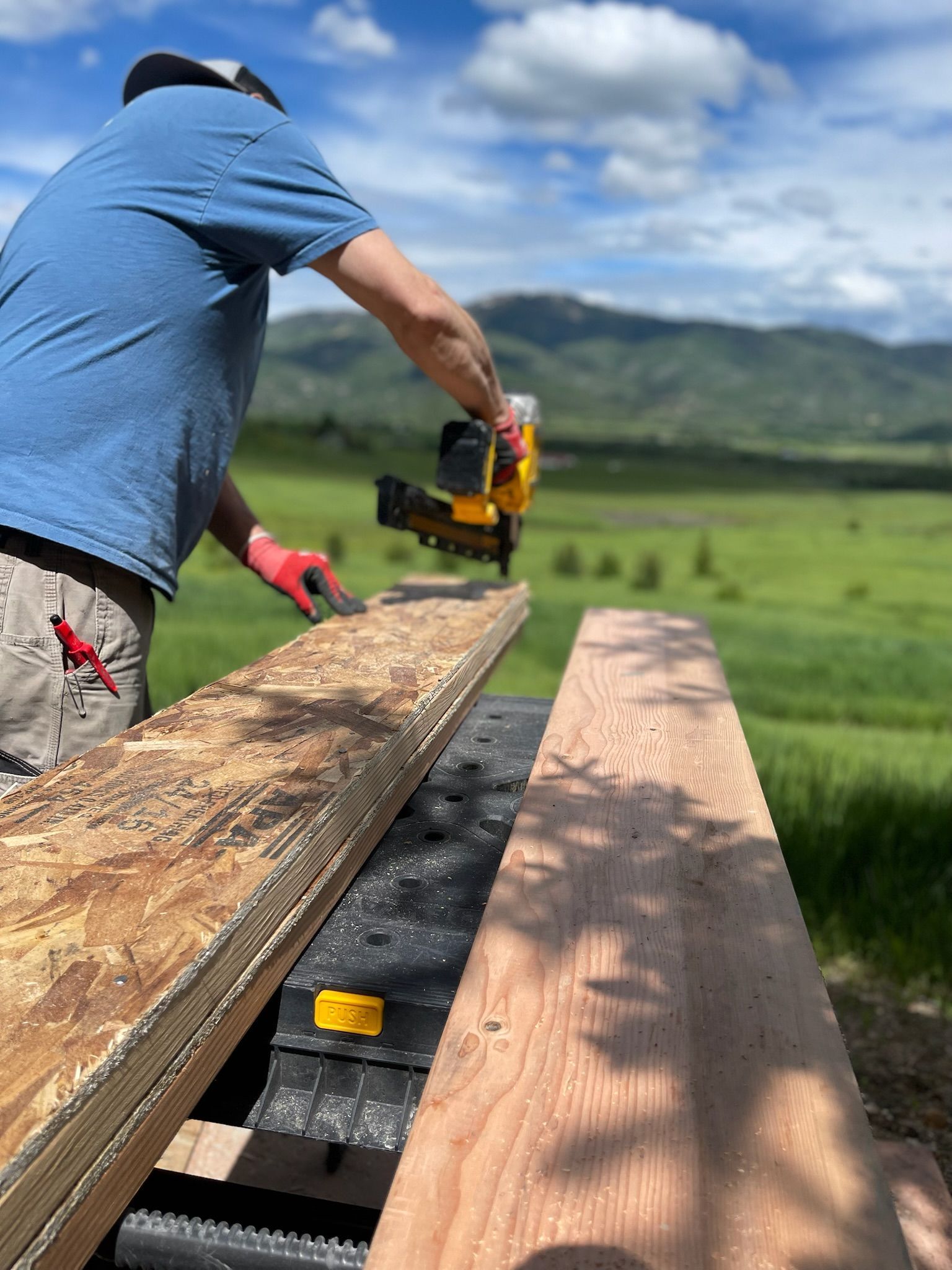 Person using a nail gun on wood outdoors, with a mountain and field backdrop.