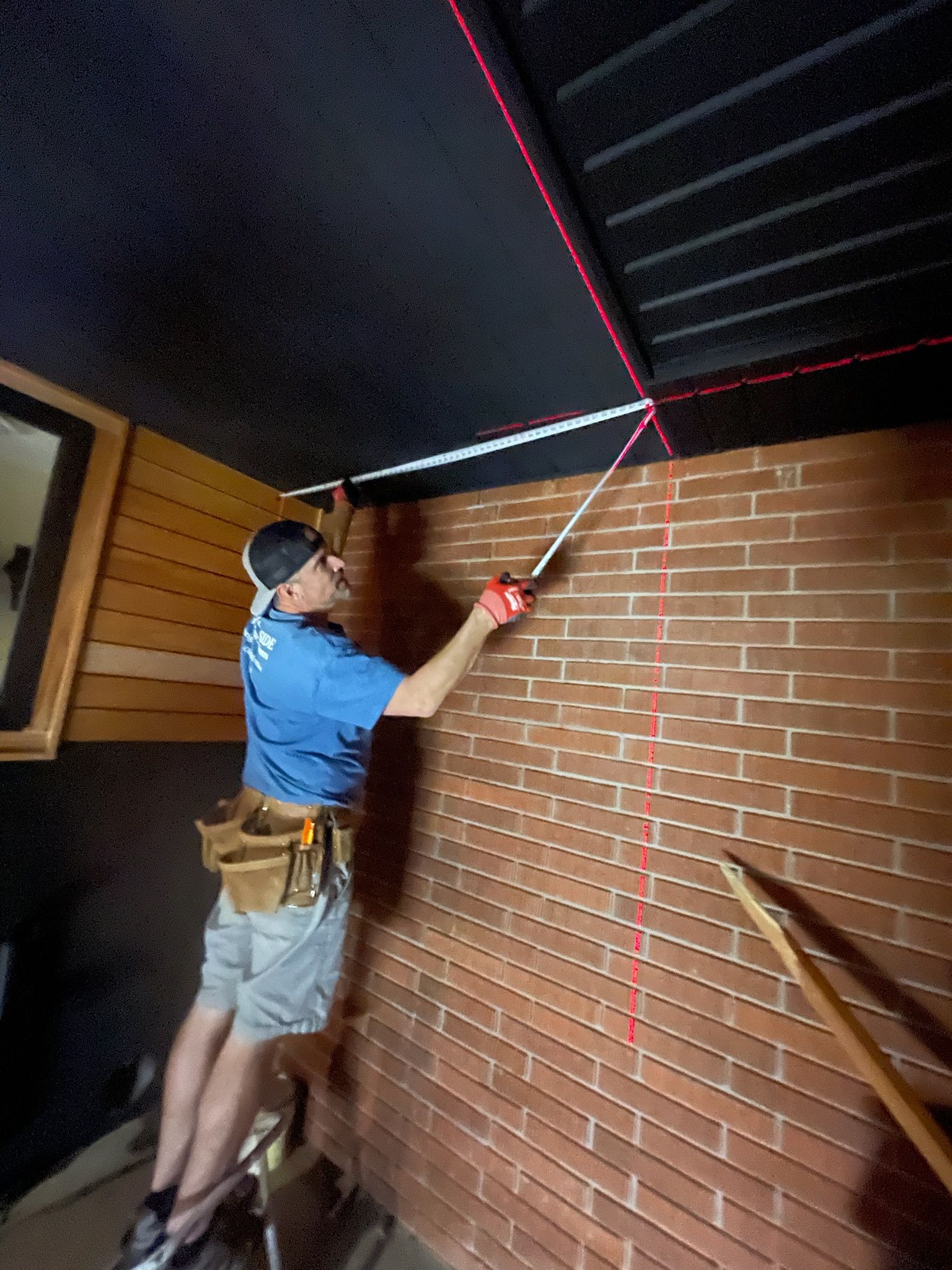 Person using a laser level and measuring tape on a brick wall, red laser lines on wall and ceiling.