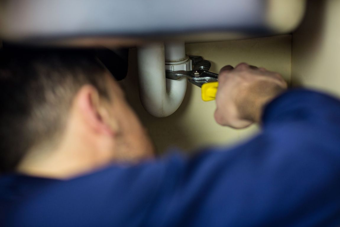 A plumber in a blue shirt tightens a PVC pipe under the sink.