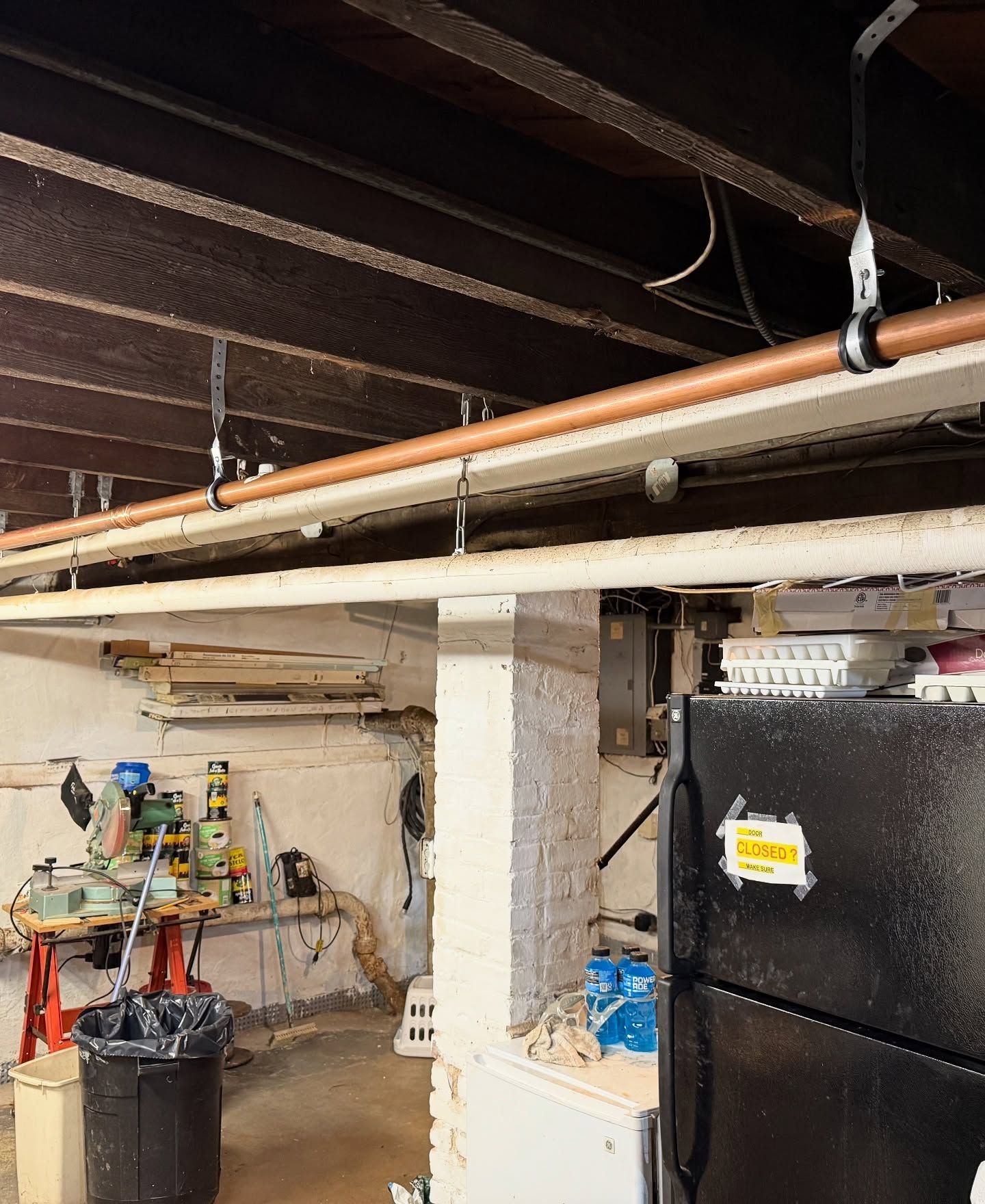Basement interior with copper pipe, old ceiling beams, white brick support, black refrigerator.