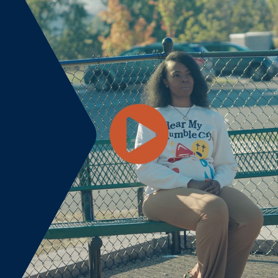 Woman seated on a park bench, wearing a sweatshirt, looking thoughtful. Sunny outdoor setting, fence, and trees.
