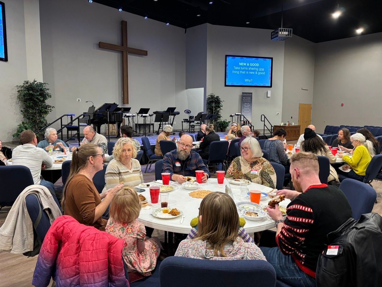 People seated at round tables in a church hall, eating, with a cross and stage in the background.