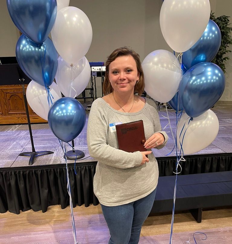 Woman holding a book, smiling, surrounded by blue and white balloons on a stage.