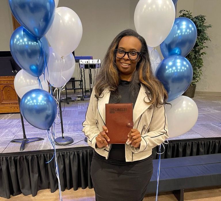 Woman in glasses holds a book in front of blue and white balloons. She is in a stage setting.