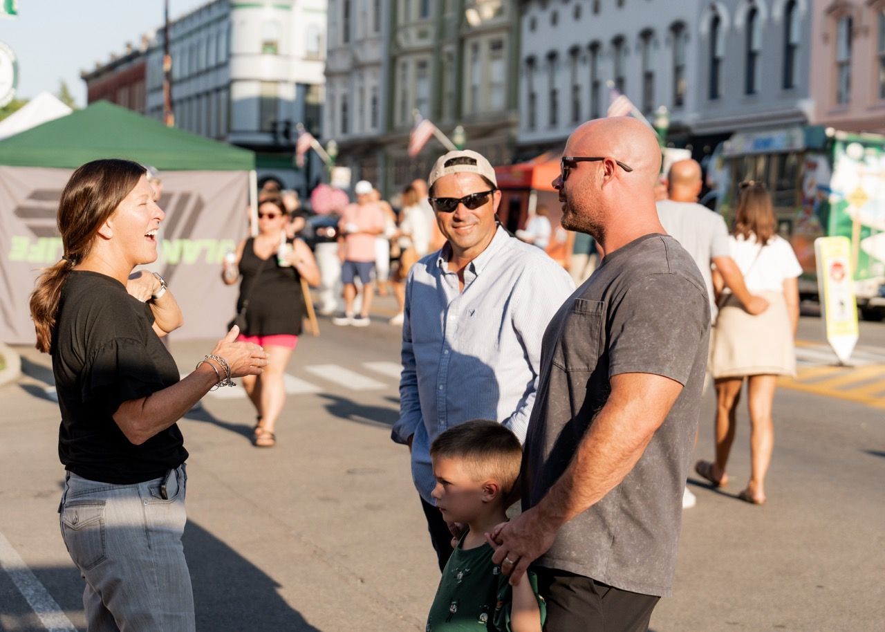 People talking on a street fair; woman gesturing; man with son; buildings in background.