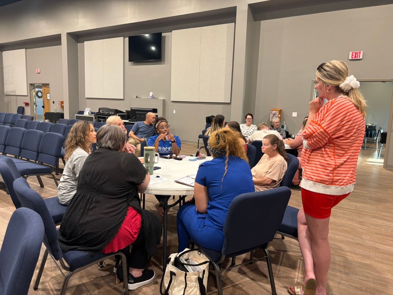 A group of people seated at a round table in a large room with a woman standing and speaking.