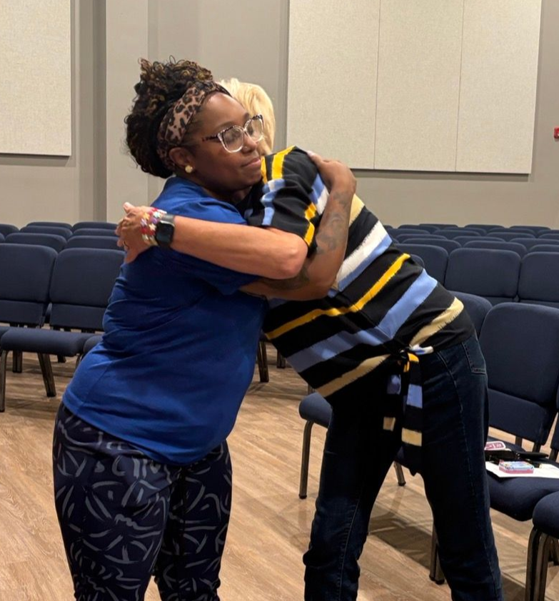 Two people embracing in a room with rows of blue chairs. Woman in blue hugs person in striped shirt.