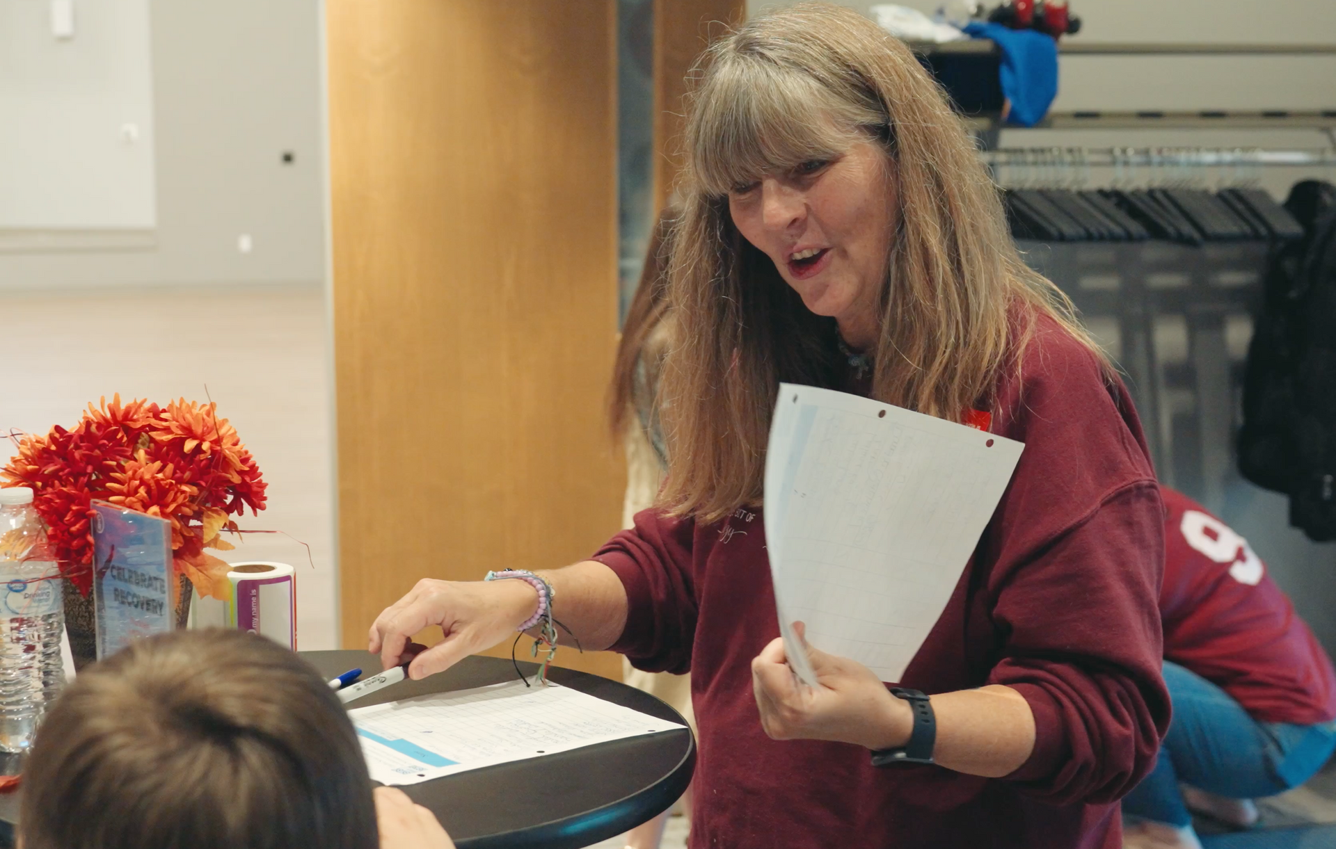 Woman in maroon top talking, holding papers, pointing at a clipboard on a table in a lobby.