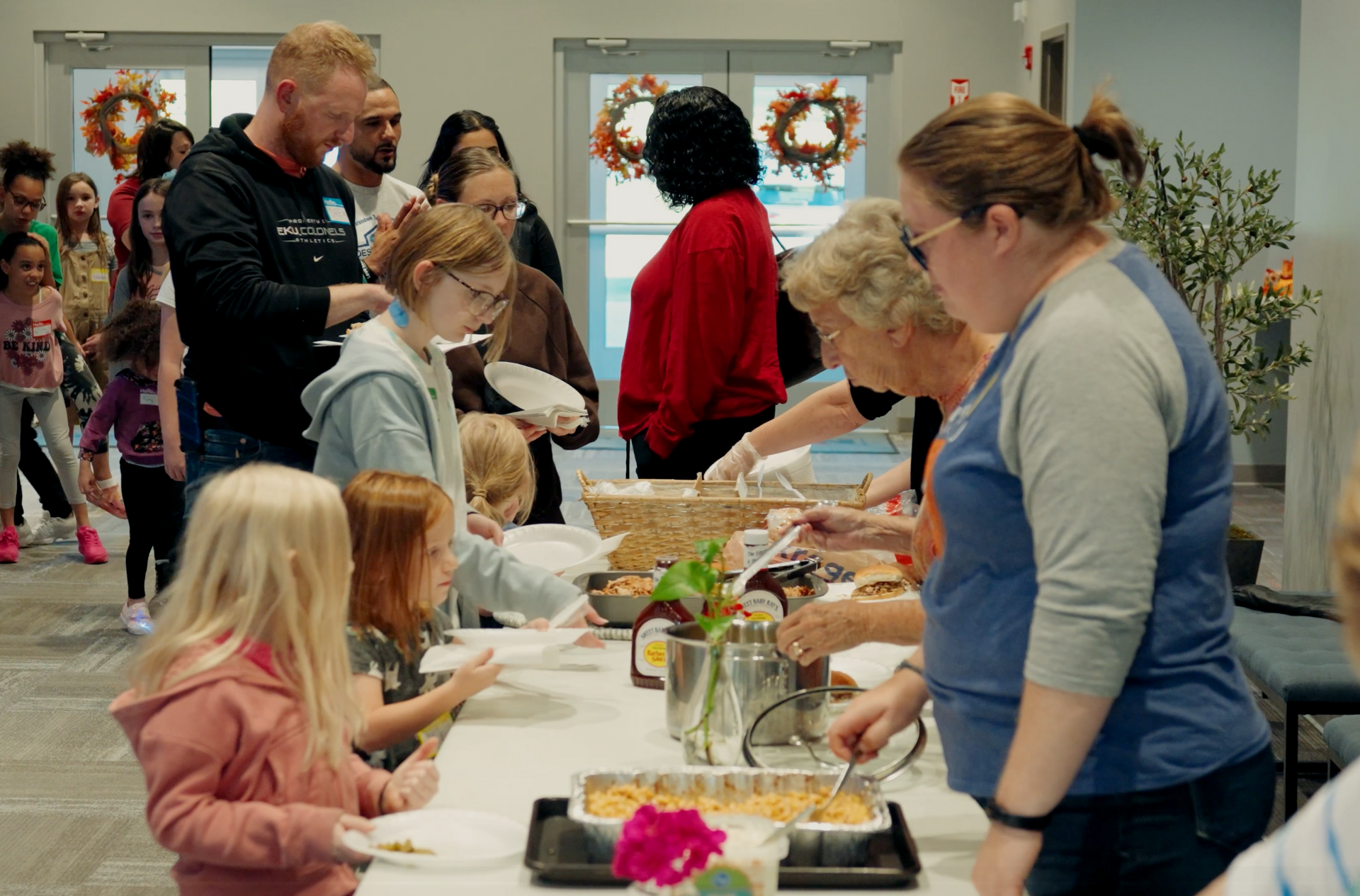 People at a food serving line, some are children. Serving table has casseroles and sides.