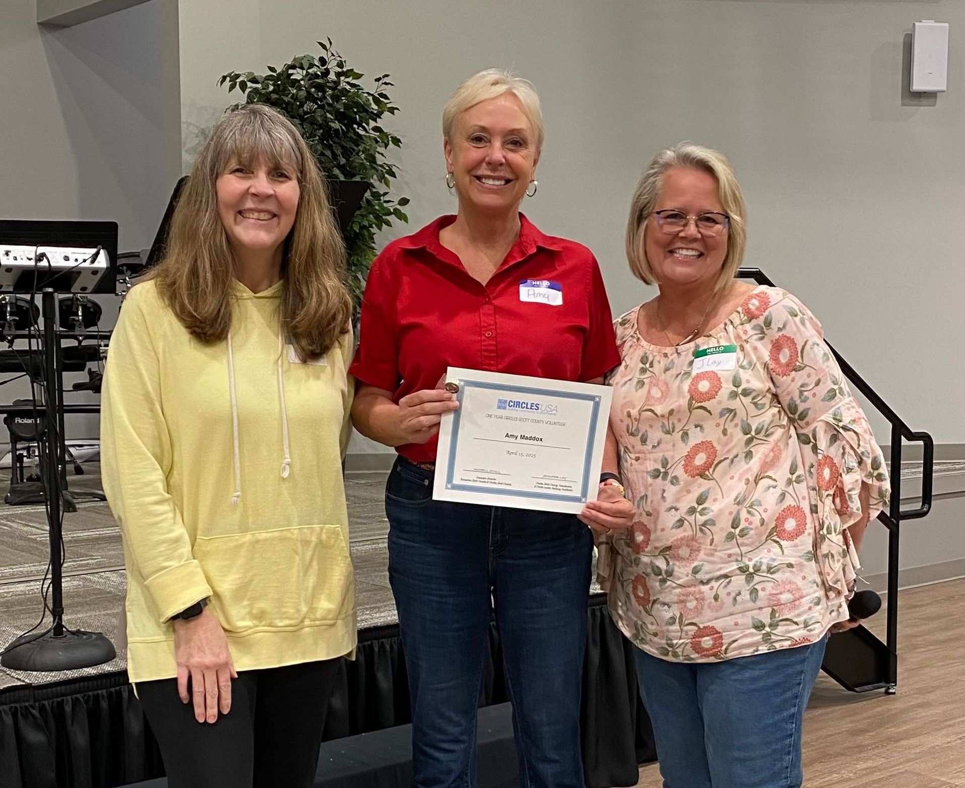 Three women smile while holding a certificate. One wears a red shirt, two are in casual clothes, at an event.