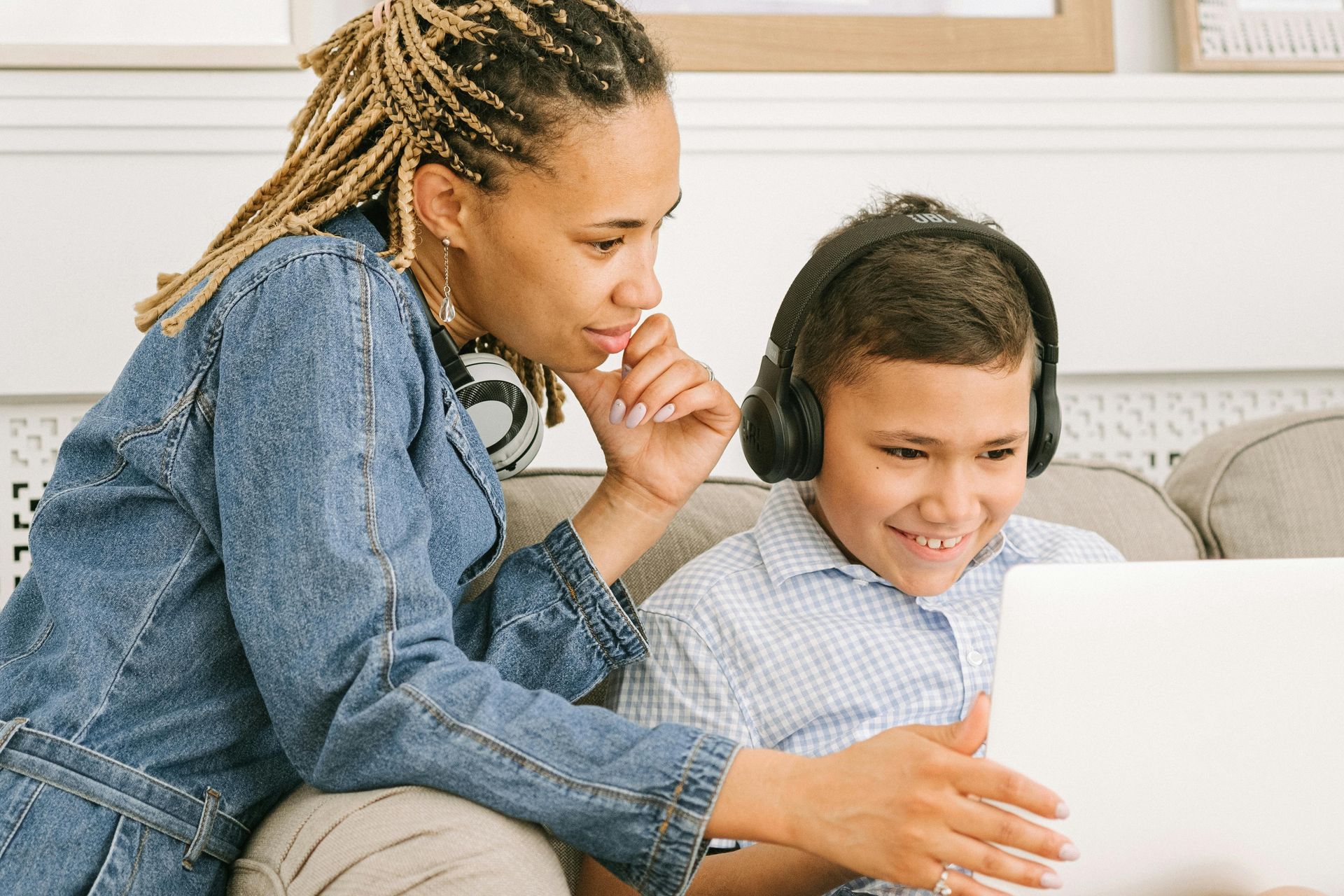 Woman in denim jacket and boy with headphones look at a laptop, both smiling.