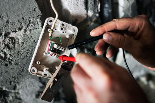 Hands testing electrical wiring in an open junction box on a gray wall.