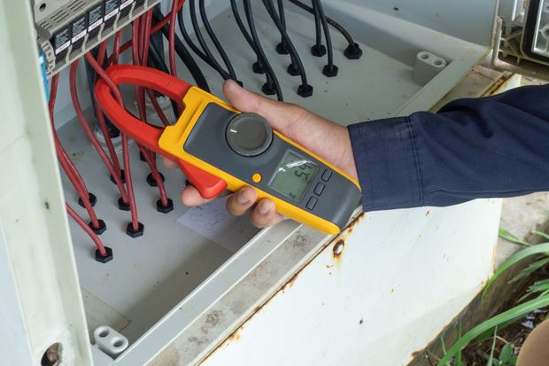 A person using a yellow clamp meter to measure the current of electrical wires inside a gray electrical box.