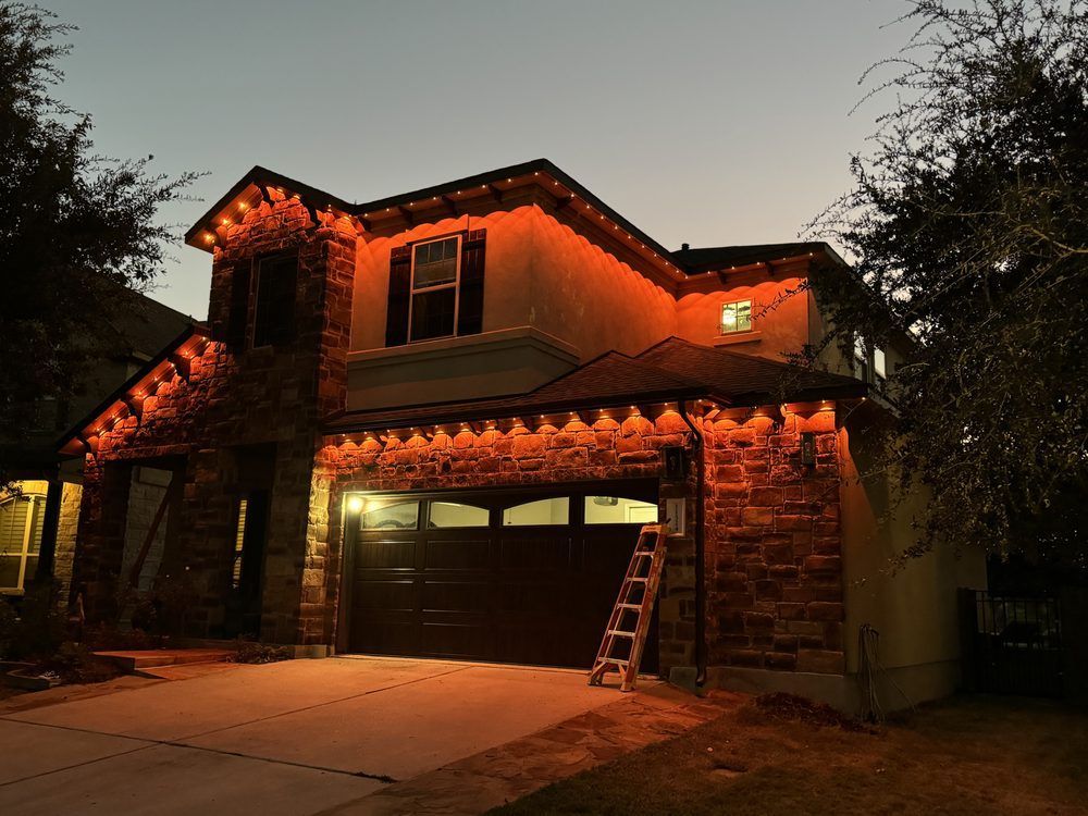 House with orange lights along the roofline, lit at dusk; ladder propped against the garage door.