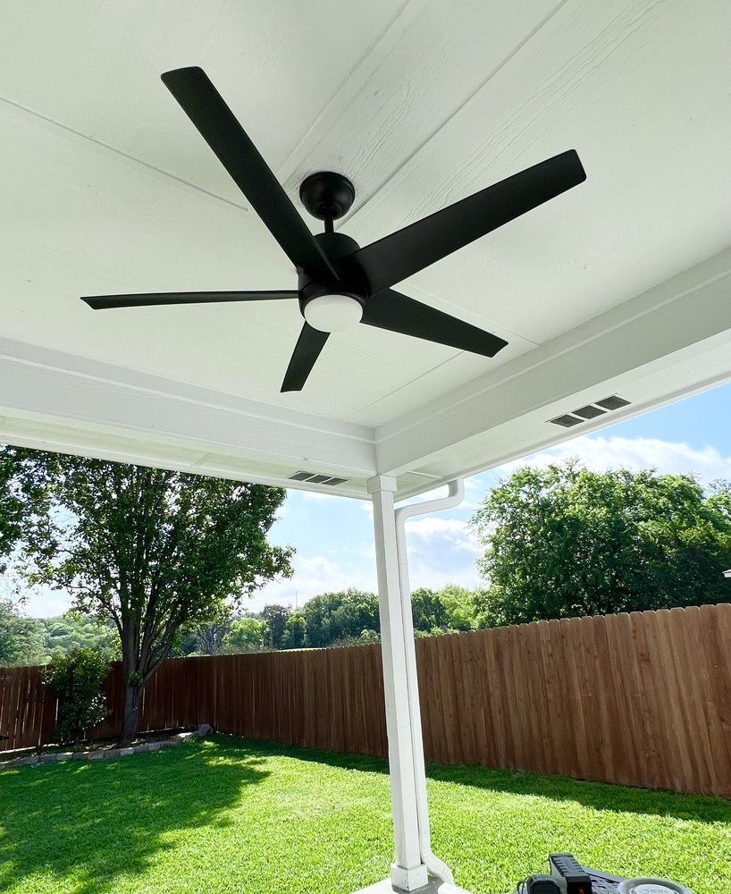 Black ceiling fan with five blades, mounted on a white covered patio, overlooking a green lawn and wooden fence.