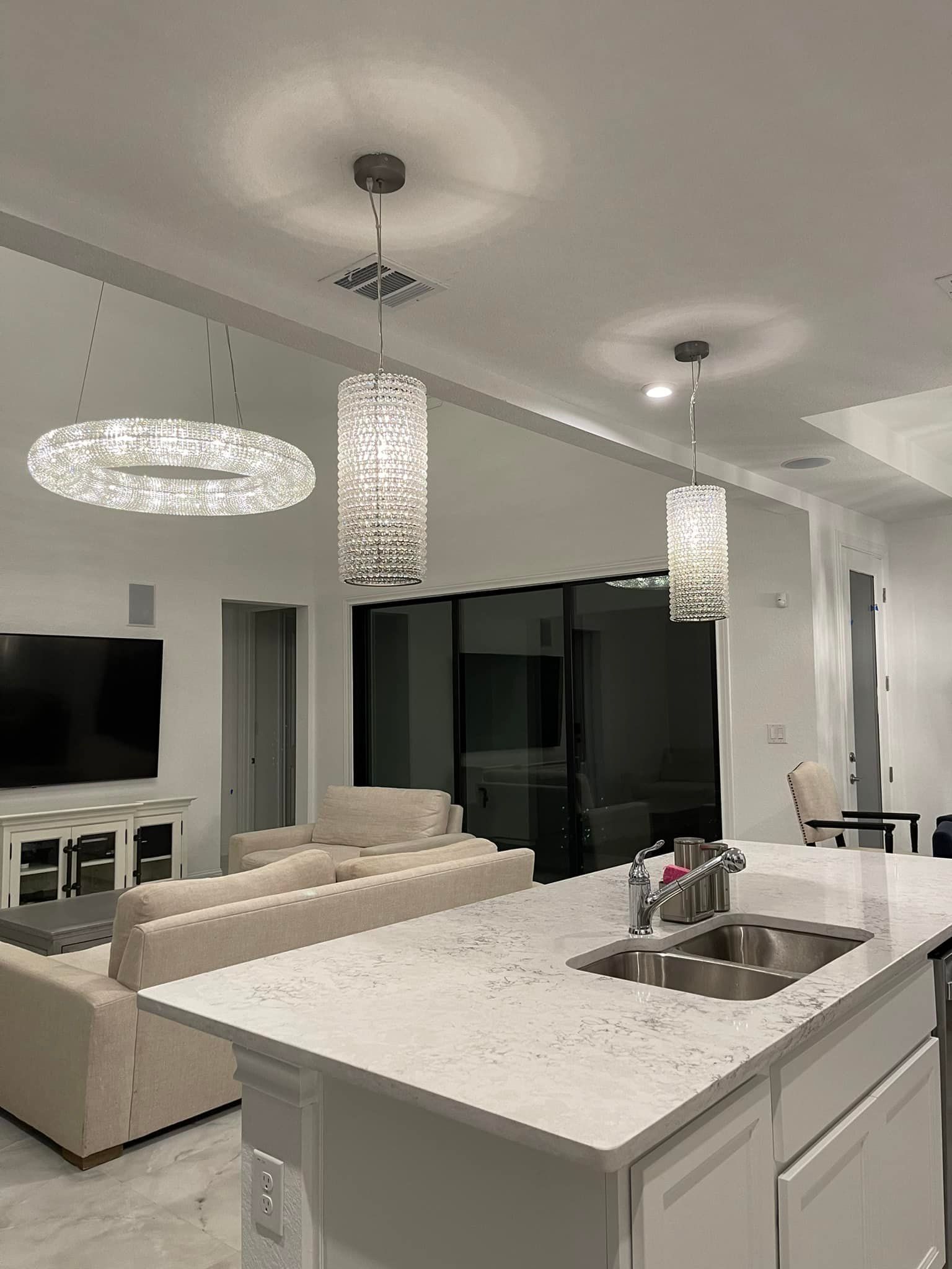 Modern kitchen with white cabinets, island, and pendant lights. Living room with a couch and TV visible.