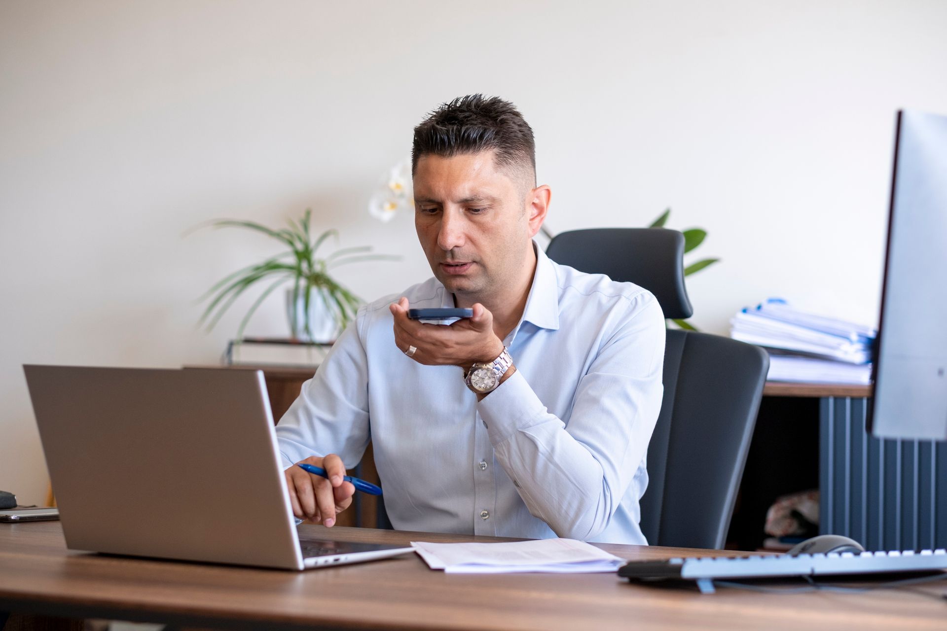 A man is sitting at a desk using a laptop and talking on a cell phone.