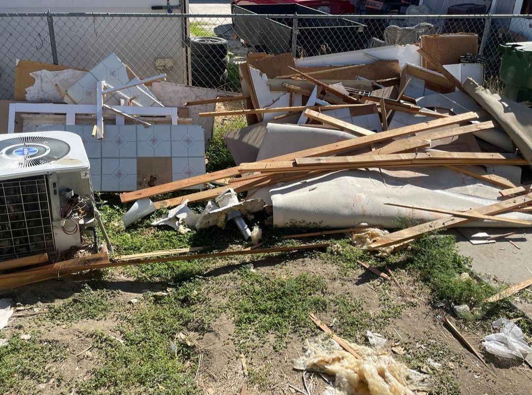 Pile of construction debris and an air conditioning unit on grass, near a chain-link fence.