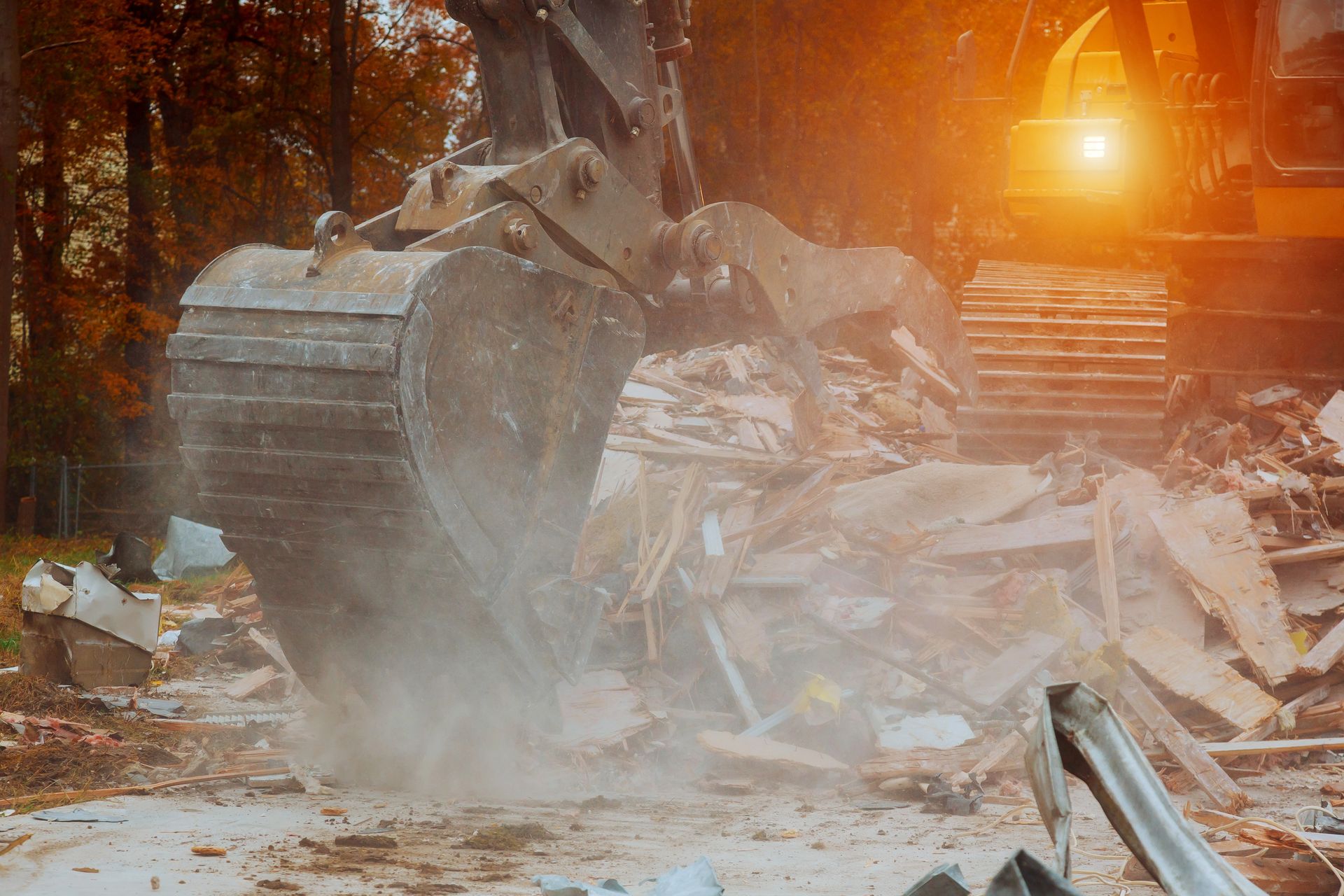 Excavator demolishing a structure, kicking up dust; bright light in the background.