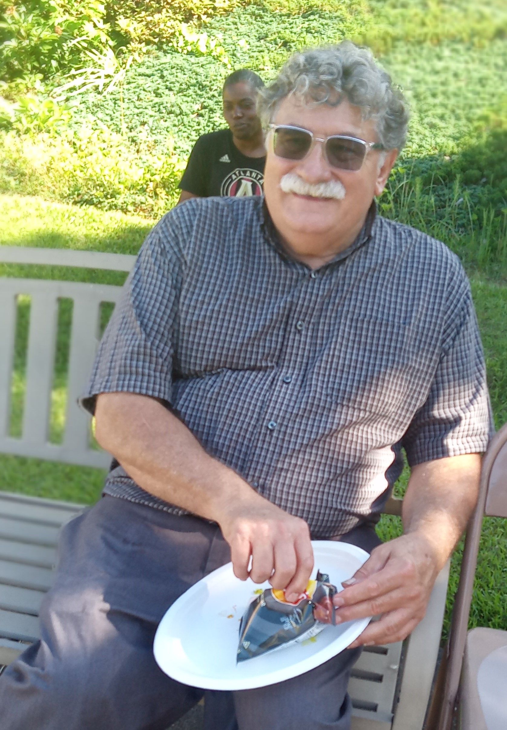 A man with a mustache is sitting on a bench holding a plate of food.