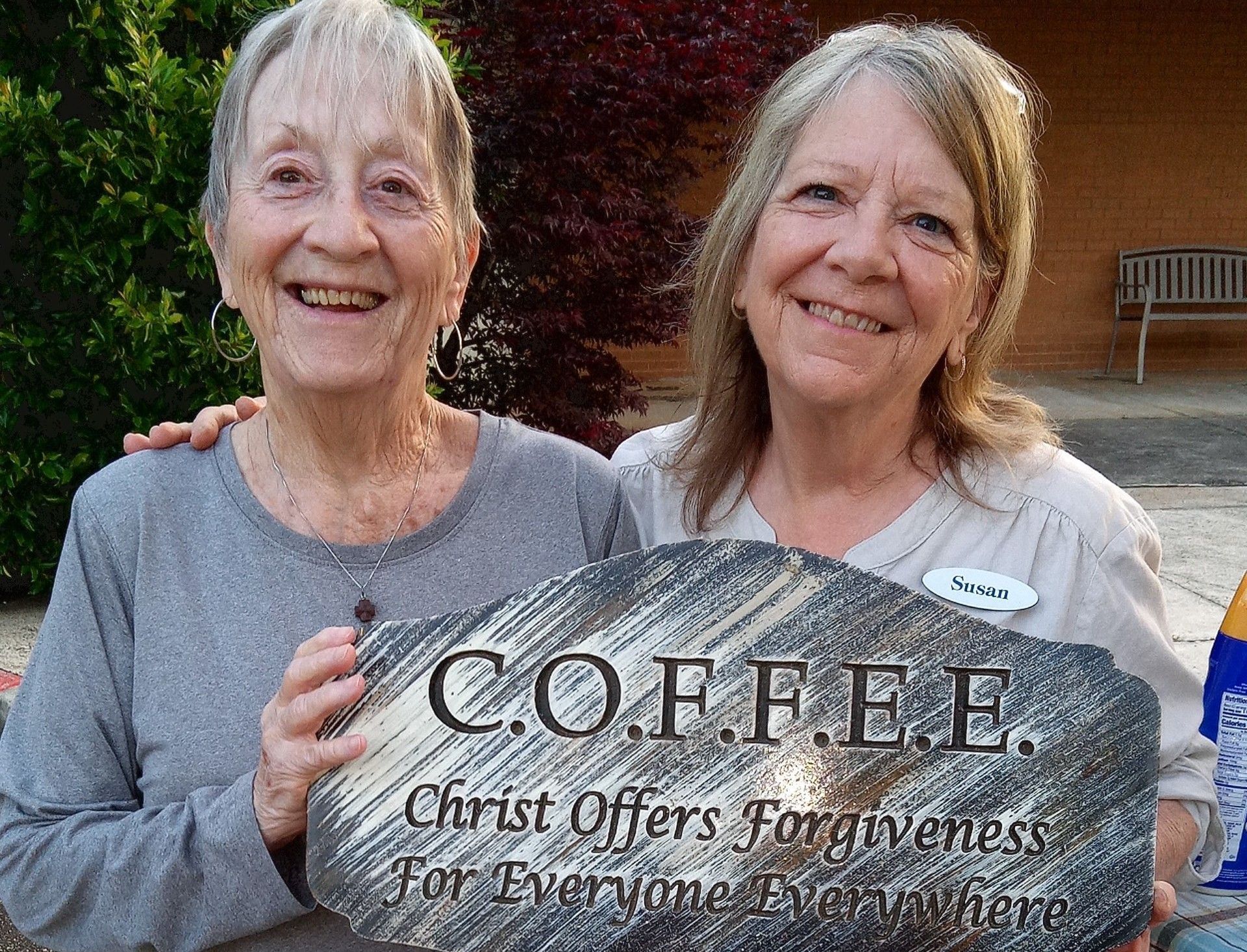 Two women holding a sign that says coffee christ offers forgiveness for everyone