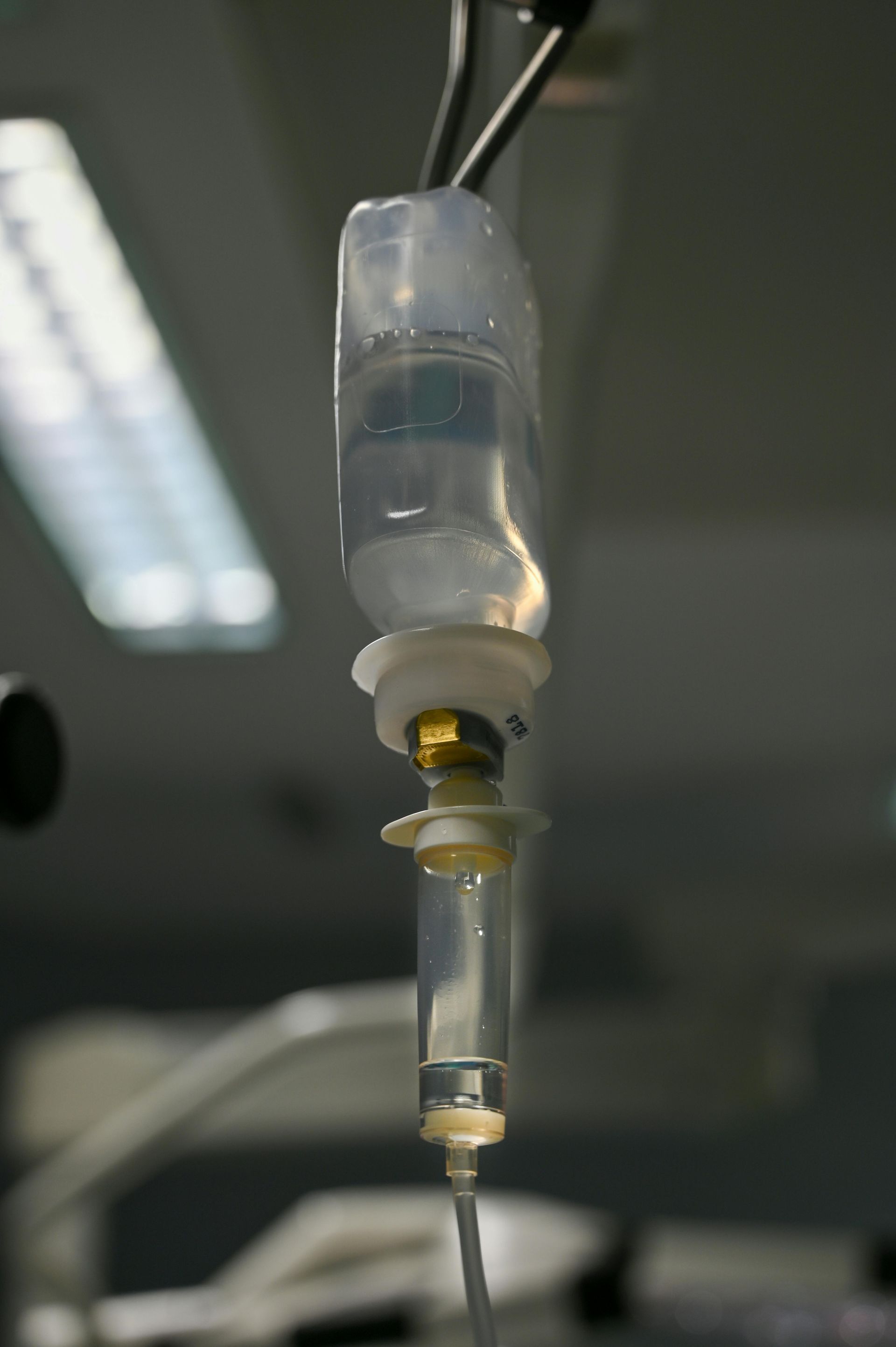 An intravenous fluid bag and drip chamber hanging against a blurred clinical background.