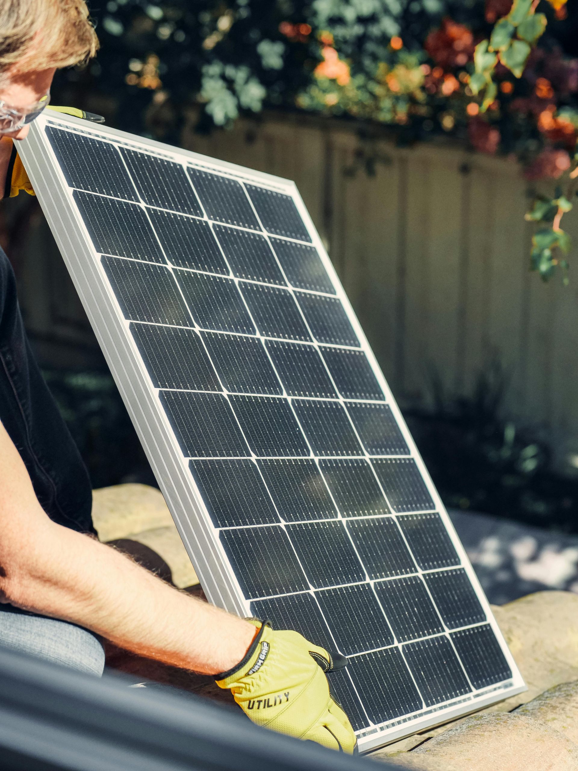 Person holding a solar panel outdoors near a fence and foliage.