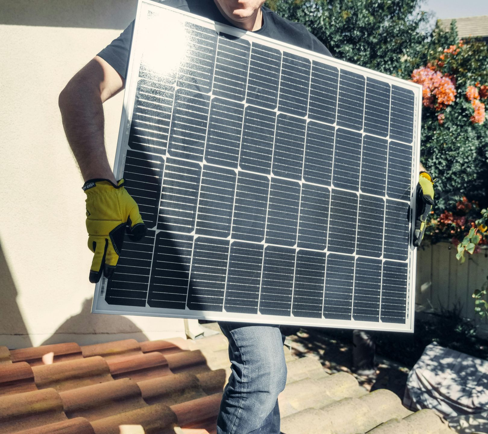 Person carrying solar panel on a rooftop near shrubs.