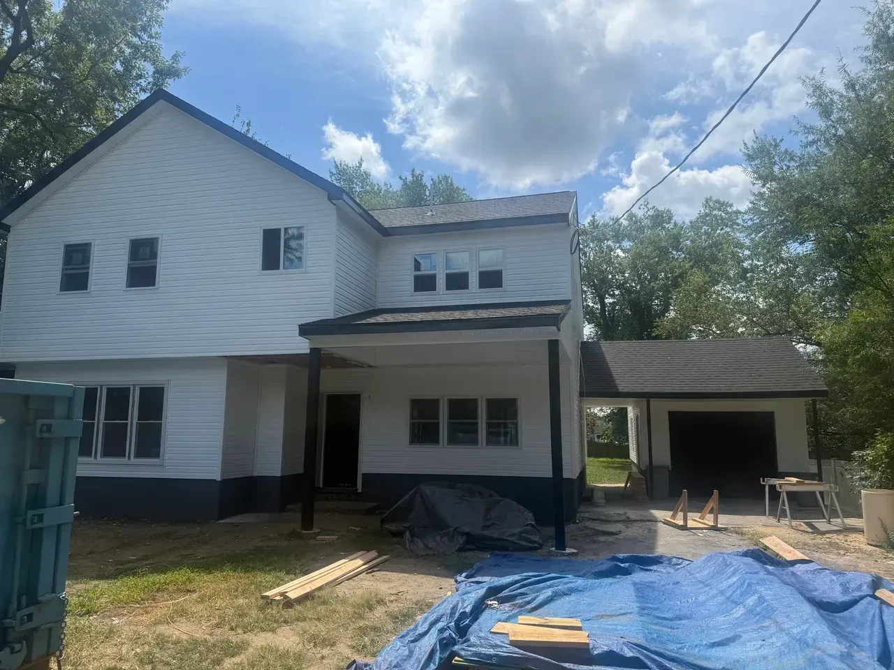 Two-story white house under renovation with blue tarp, porch, and detached garage in the yard.