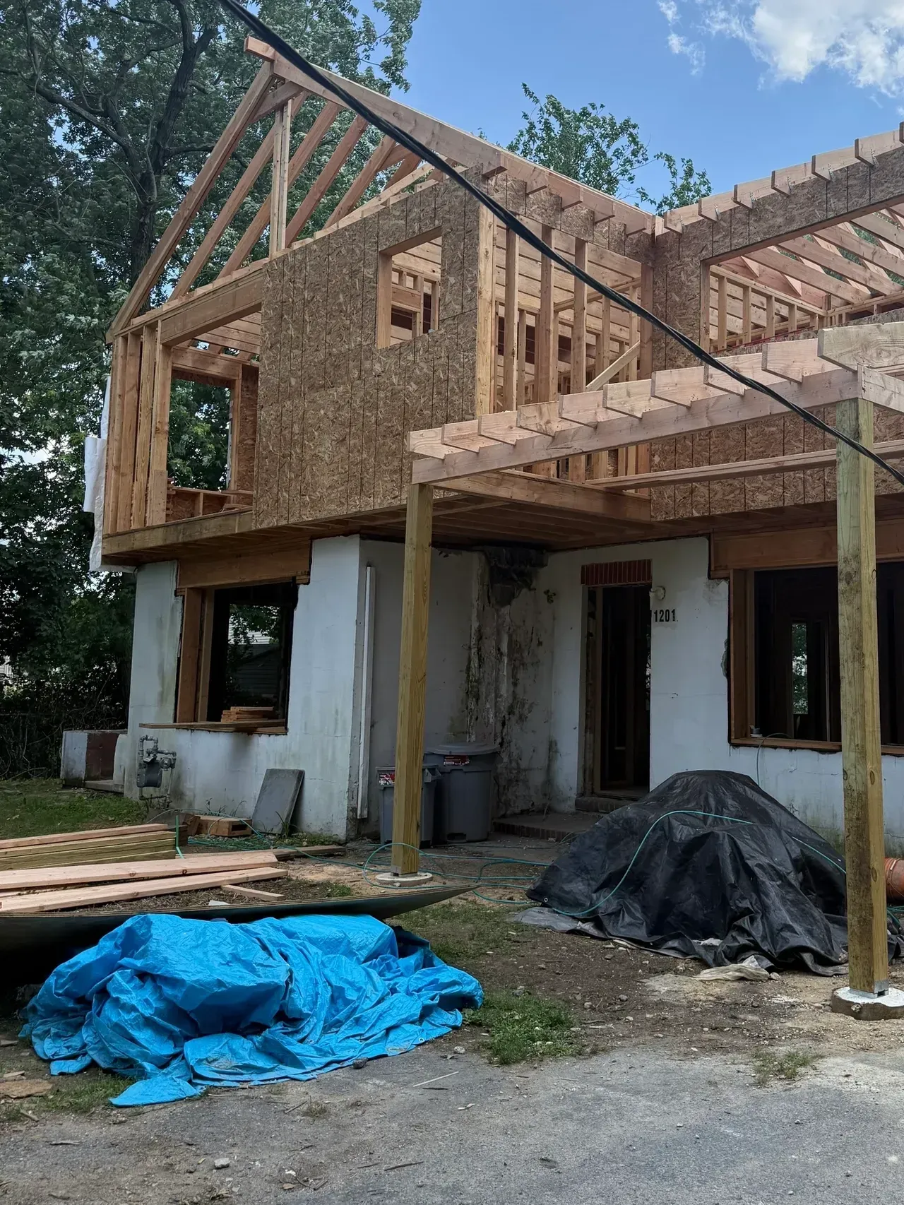 Partially renovated house with exposed wooden framing, blue tarp, and construction materials in front yard