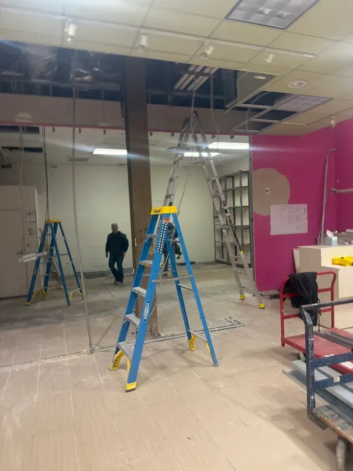 Office space under renovation with ladders, scaffolding, and a worker in a partially removed ceiling