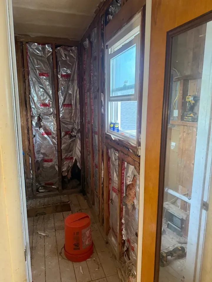 Narrow unfinished hallway with exposed insulation, wood framing, a window, and an orange bucket on the floor