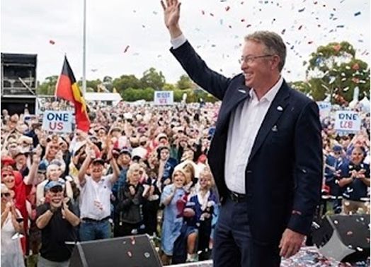 Man in suit waves to a cheering crowd at an outdoor event with confetti and signs.