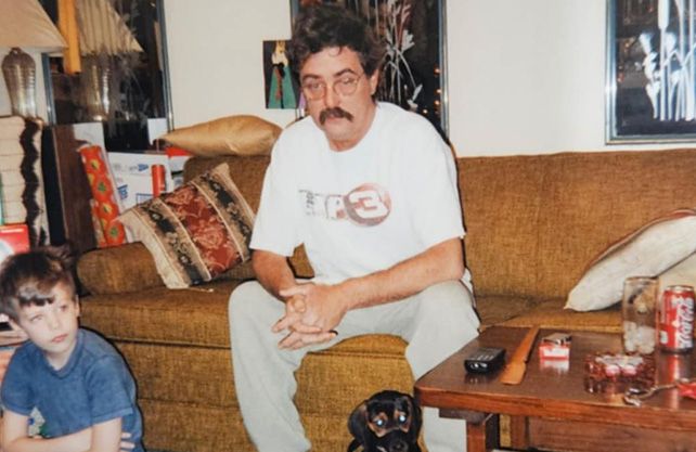 Man on brown sofa with boy and dog; small table with objects.
