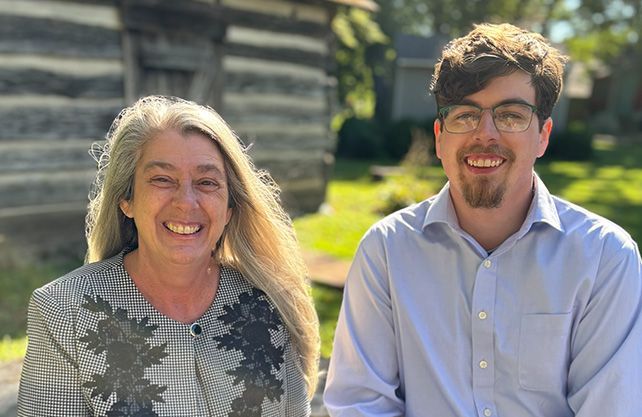 Two people smiling outdoors, in front of a wood building.