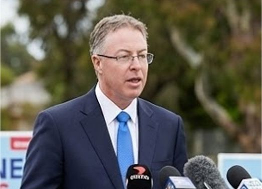 Man in a blue suit speaks at a press conference outdoors, with microphones in front of him.