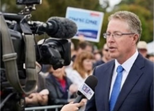Man in suit being interviewed by a reporter with a camera. People and a sign are in the background.