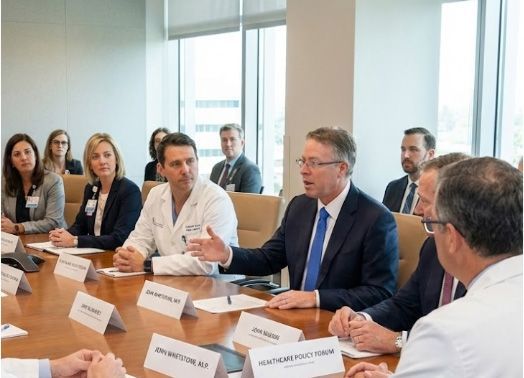 Group of people in a meeting at a conference table with nameplates. Man in suit speaks; others listen.