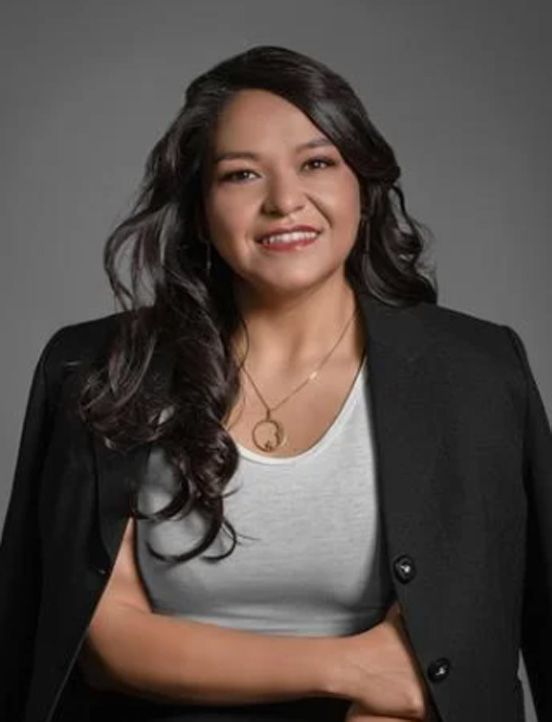 Woman in black blazer over white top, smiling, necklace, dark wavy hair.