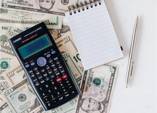 Calculator, notepad, and pen on top of scattered US dollar bills.