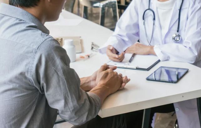 Doctor in white coat consults with patient seated at a table, discussing health.