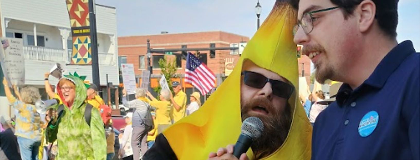 Man in banana costume interviewed, crowd with signs in background, sunny day.