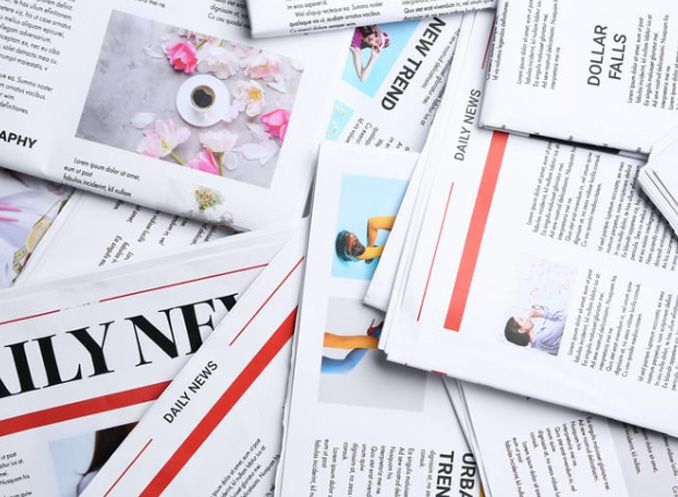Pile of newspapers, close up. Sections feature photos, headlines, and text. Red and black text on white pages.