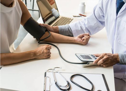 Doctor taking a patient's blood pressure with stethoscope and laptop on a desk.
