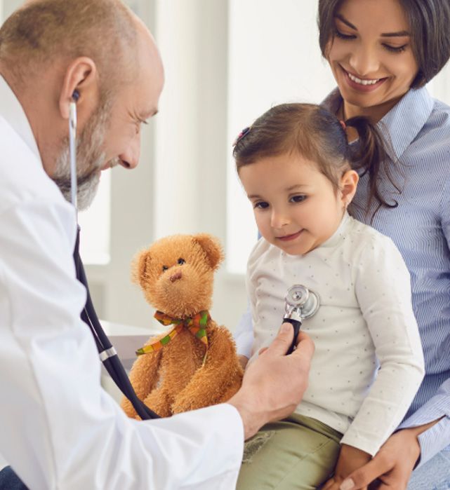 Doctor examining a child with a stethoscope while the mother watches. Teddy bear sits nearby.